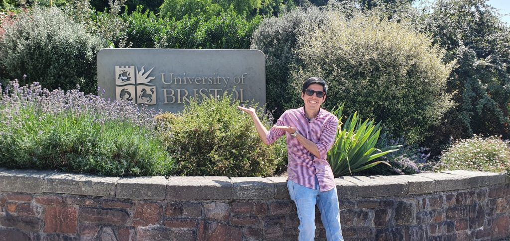 Victor Flores in a check shirt and sunglasses, gesturing happily at a sign for the University of Bristol.