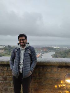 A young Inf=dian man wearing glasses and a denim jacket smiling with the Avon Gorge in the background. 