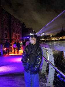 A young Thai woman in a black jacket stood smiling on a bridge, with purple lights in the background. 