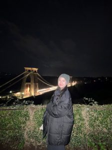 A young Thai woman in a black coat and grey beanie standing in front of the Clifton Suspension Bridge.